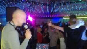 tomislav 3 DJ Tomislav (Tom Black) drži mikrofon dok vodi program na vjenčanju, s osmijehom na licu ispred DJ pulta.